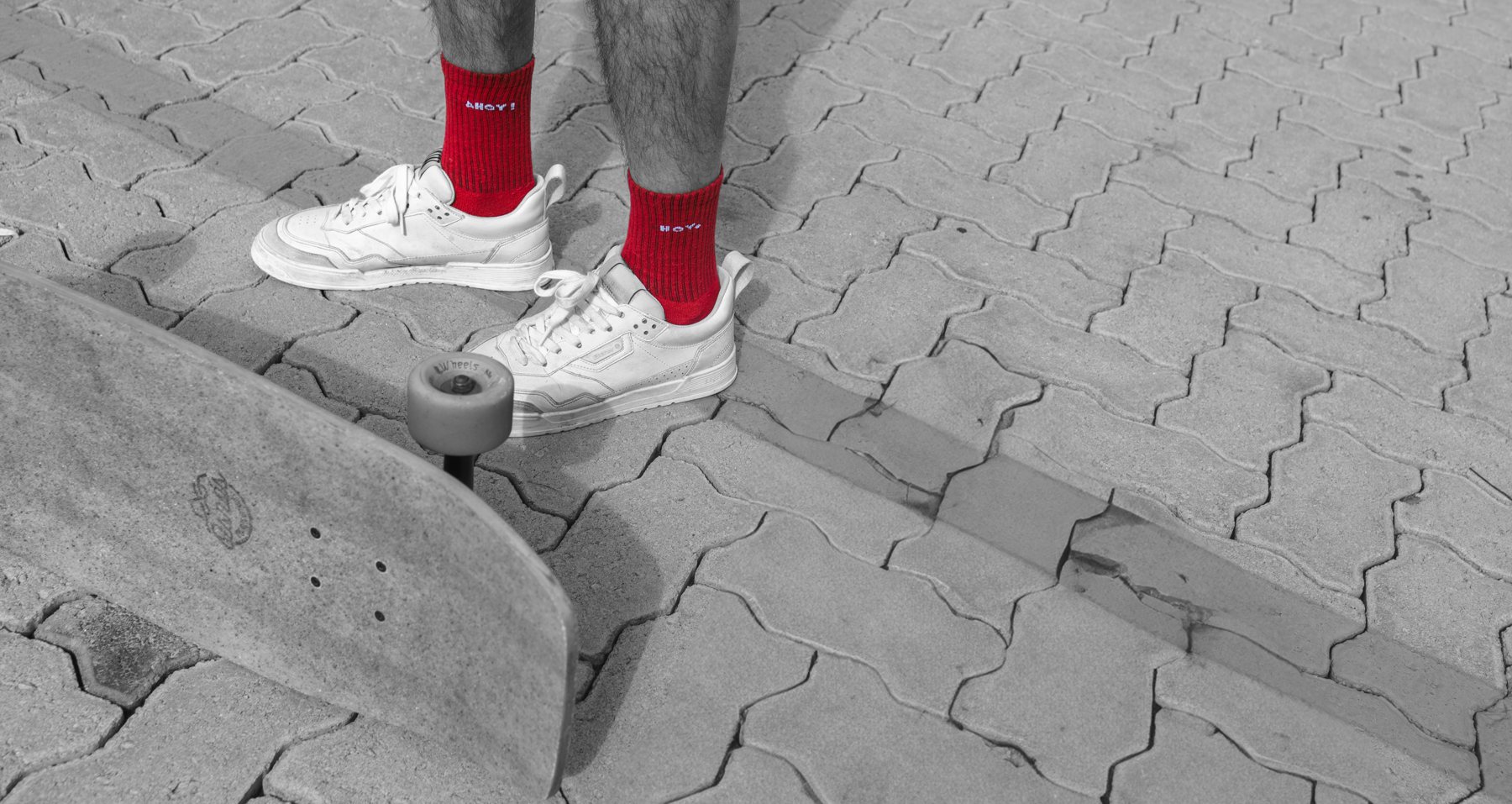 Pés com meias vermelhas Ahoy e tênis brancos ao lado de um skate, foto em preto e branco com a meia colorida